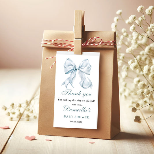Brown paper bag with a white tag featuring a blue bow and baby shower text, surrounded by white flowers on a wooden surface.