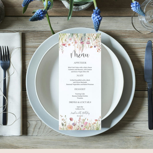 Menu card on a plate with wildflower floral design, surrounded by cutlery and flowers on a wooden table.