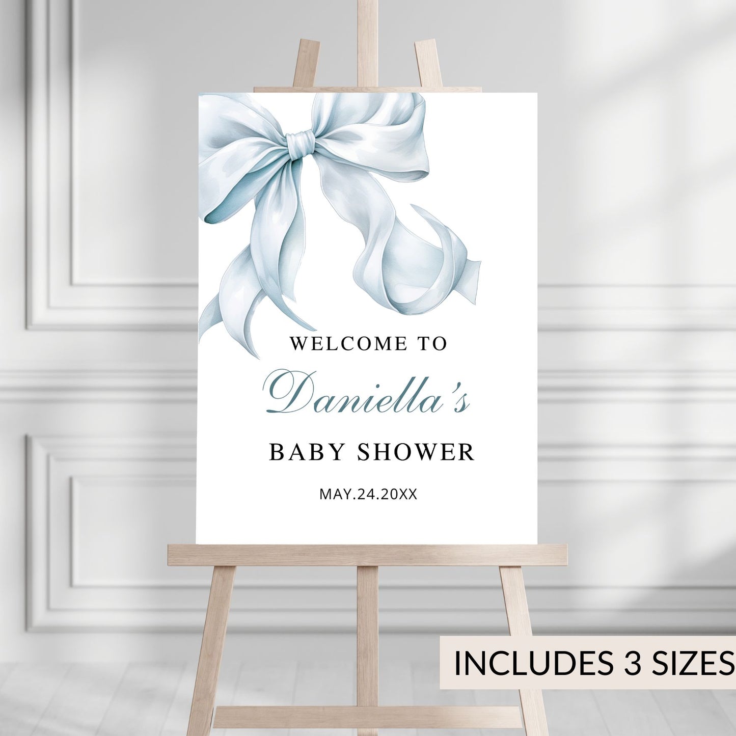 Baby shower welcome sign with a blue bow on a white background, displayed on an easel.
