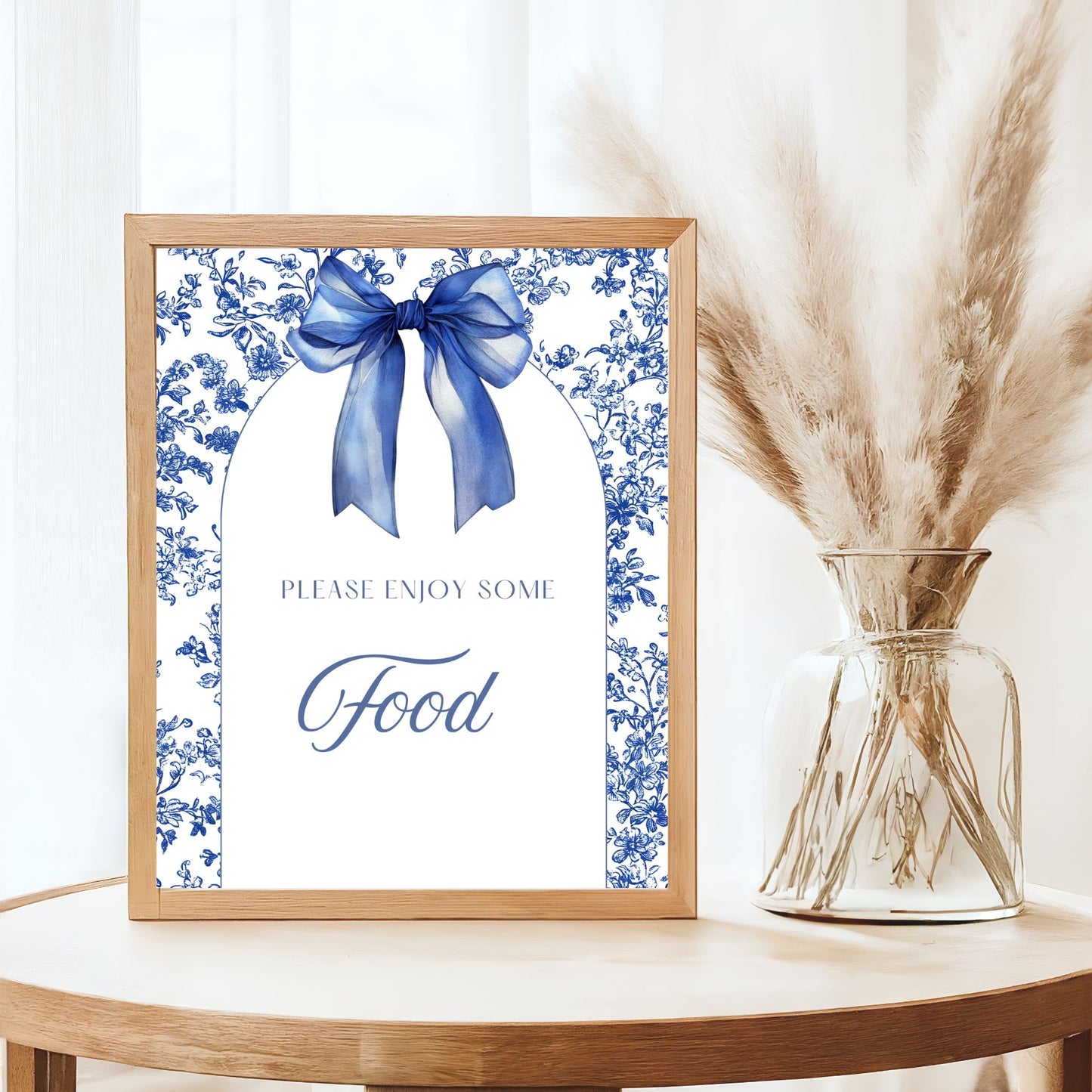Framed artwork with blue bow and floral design on a table with a vase of dried plants.