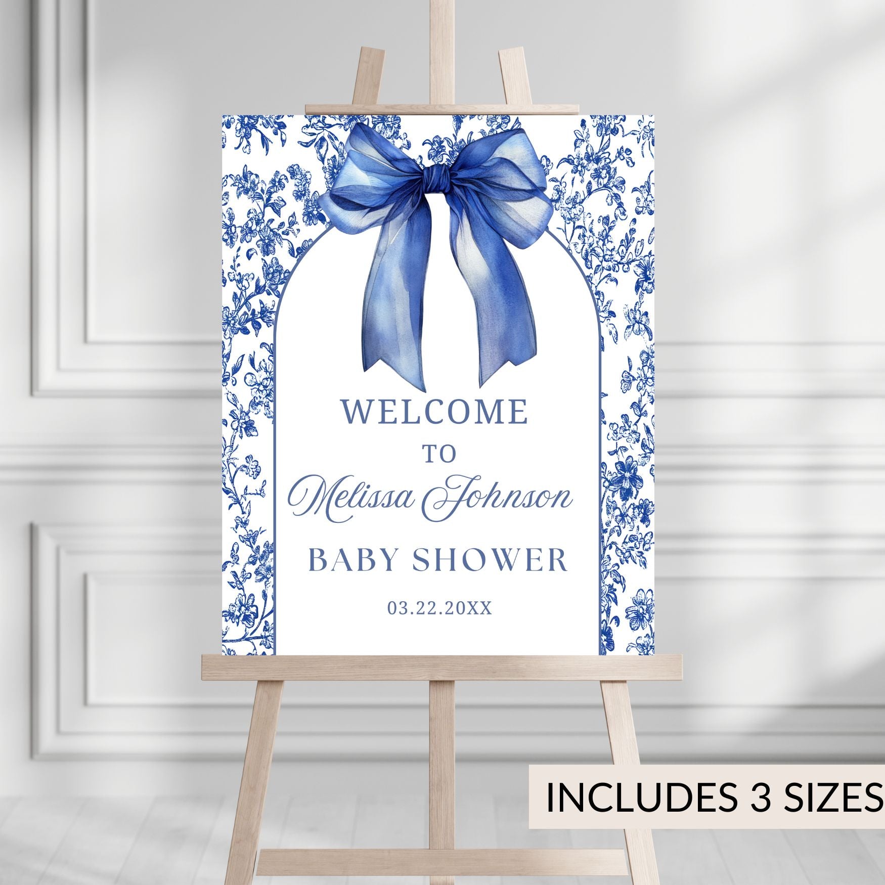 Baby shower welcome sign with blue bow and floral design on a white background, displayed on an easel.