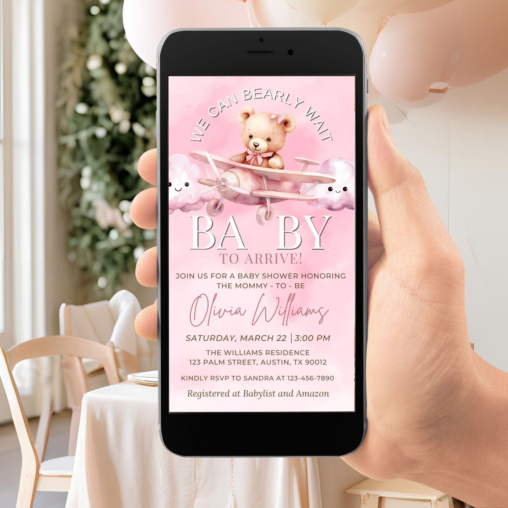 Hand holding a smartphone displaying a baby shower invitation with teddy bears and a plane on a pink background.
