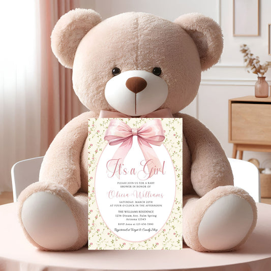 teddy bear holding a baby shower invitation with a pink bow in a room setting.