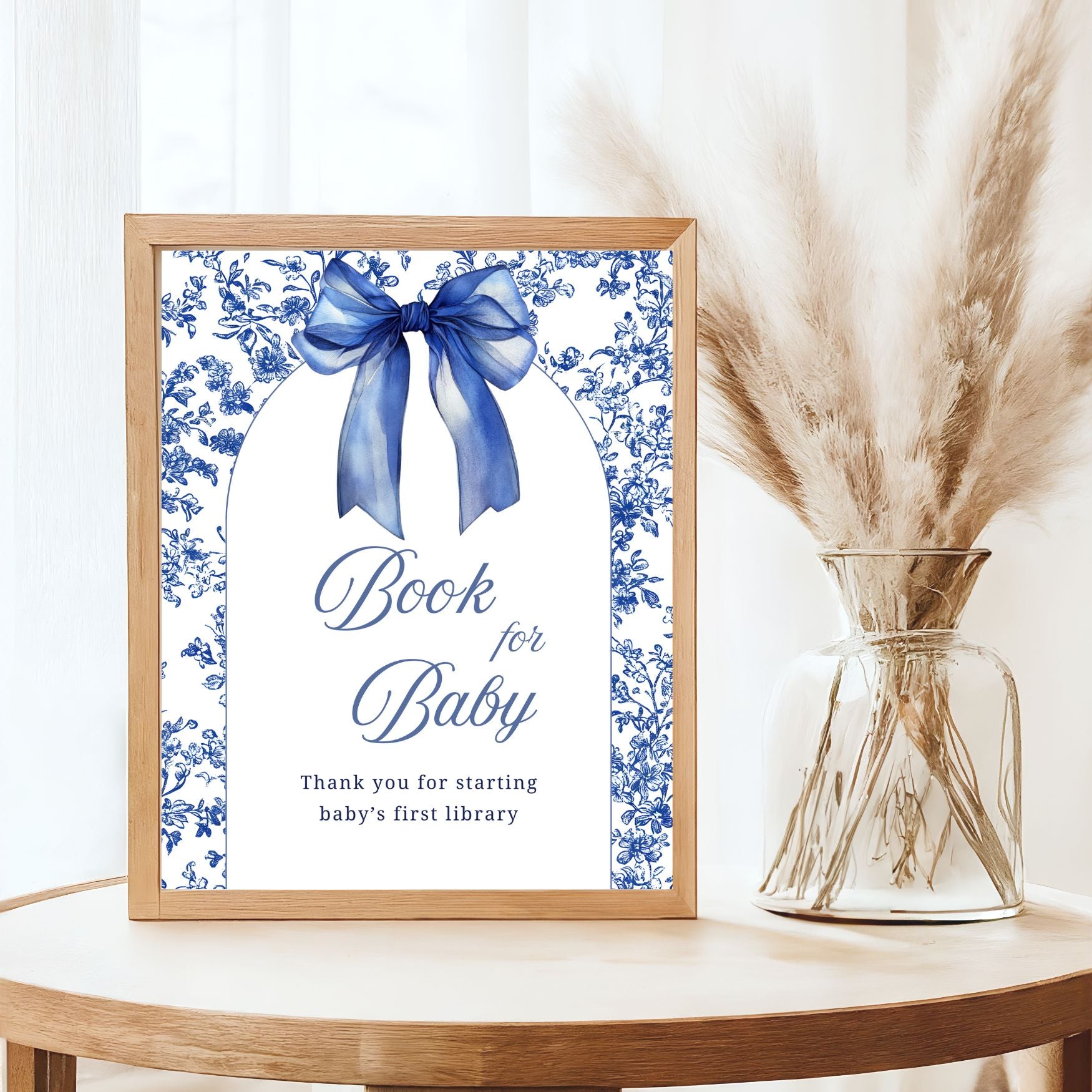 Framed artwork with blue bow and text on a white background, next to a vase with dried plants.